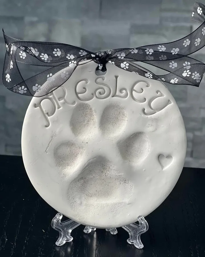 An unpainted round clay paw print with the name Presley and a small heart, on a clear stand.