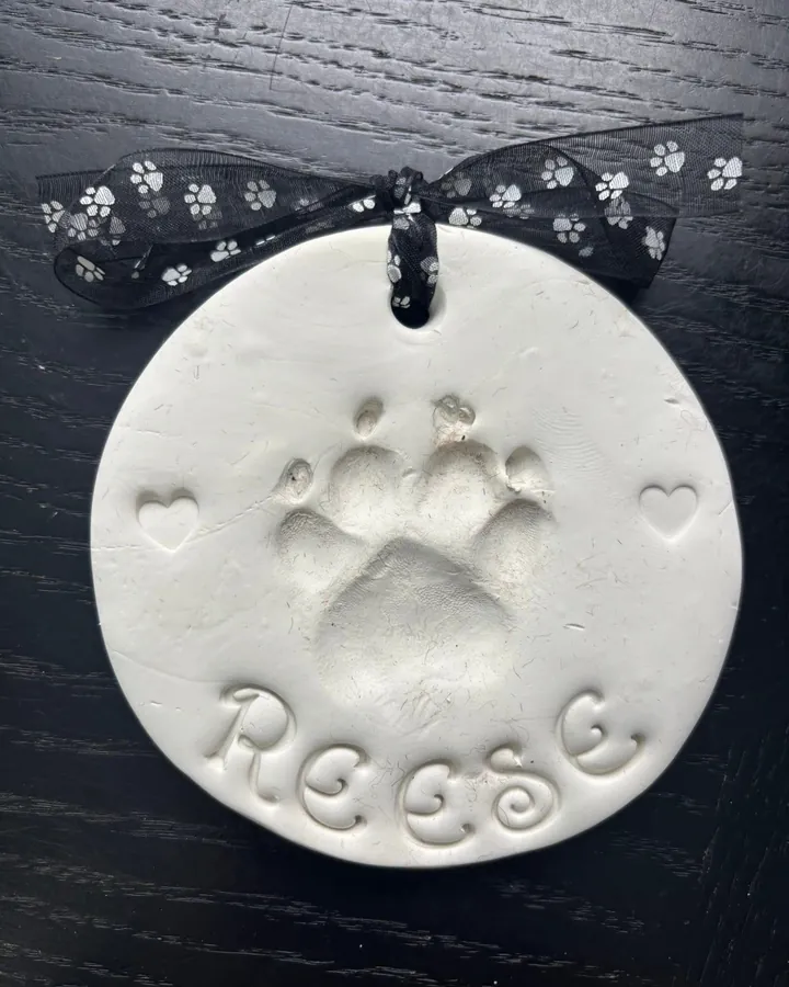 An unpainted round clay paw print with the name Reese and two small hearts, tied with a paw print ribbon.