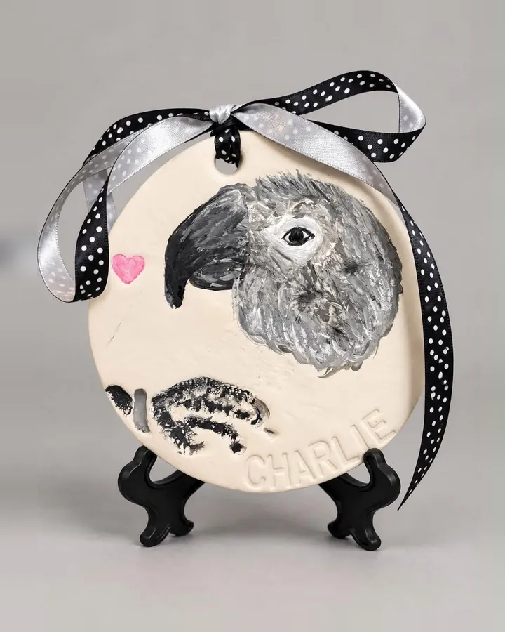 A round clay keepsake with a hand-painted portrait of a gray parrot, its painted footprint and the name Charlie, on a black stand.
