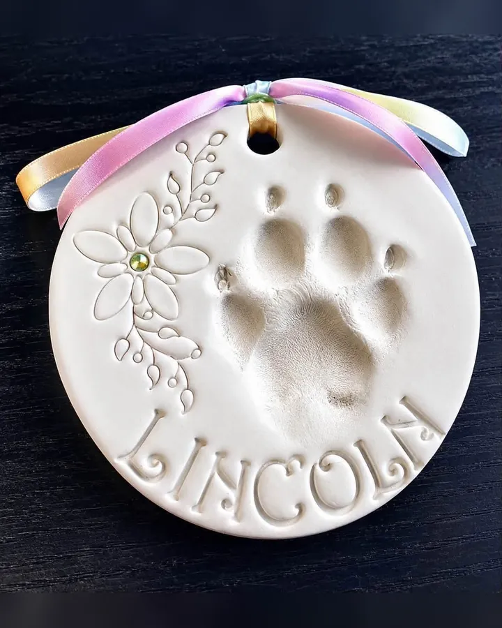 A round clay paw print with a pressed flower and leaf design and the name Lincoln, tied with a rainbow ribbon.