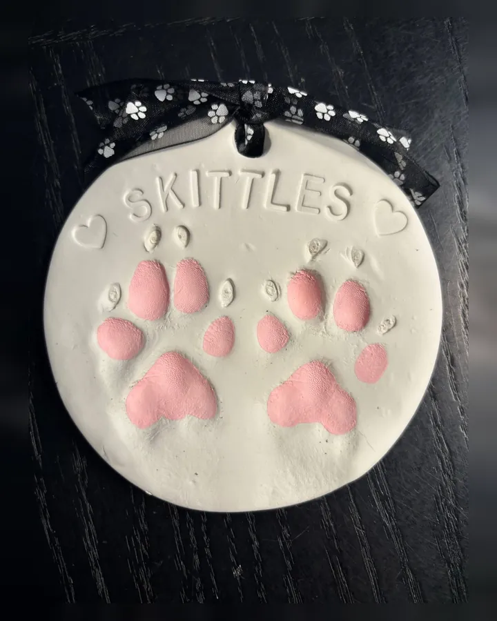A round clay print of two paws painted pink, with the name Skittles and two hearts.