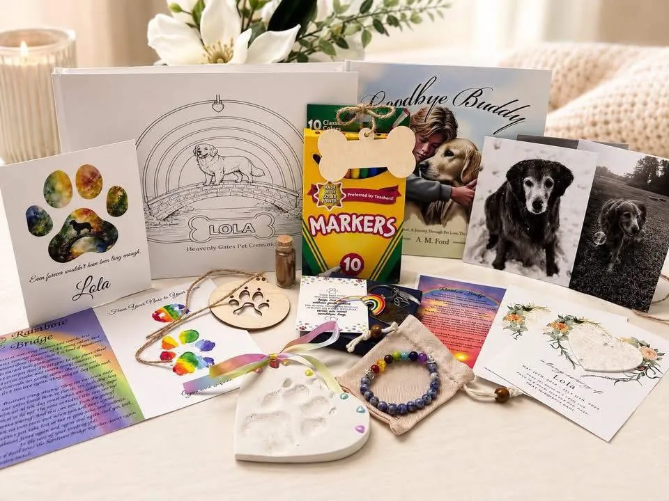 Everything from a Comfort Basket laid out on a table: the Goodbye Buddy book, a coloring page of the pet, markers, paw print cards, wooden ornaments, a heart-shaped clay paw print, a bracelet and its pouch, and photos of the dog.