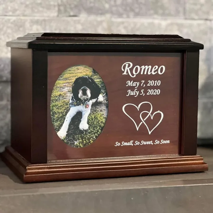 A dark wooden urn with an oval photo of a dog beside an engraved name, dates and hearts.