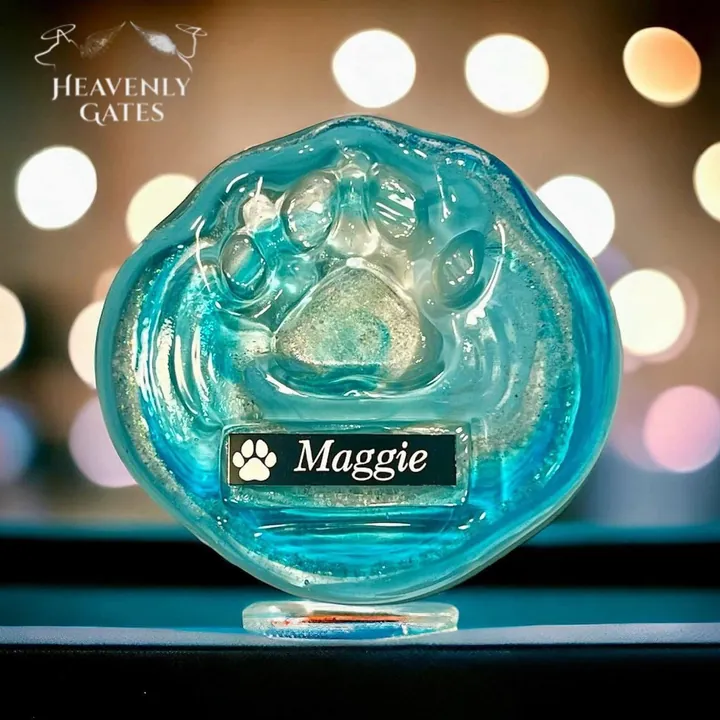 A wavy teal glass disc with a raised paw print and a black name plate reading Maggie, on a clear stand.