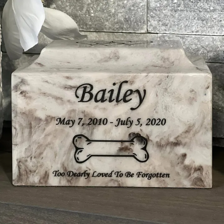 A white marble urn engraved with a name, dates, a bone and a short message.