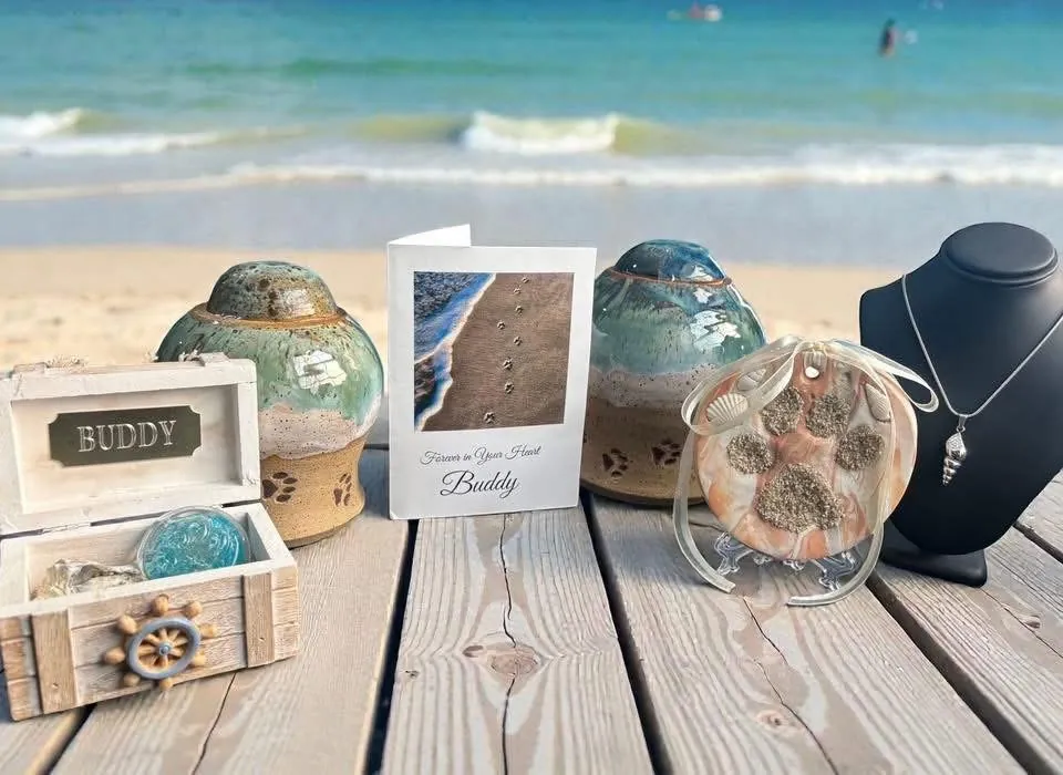 The Sandy Beach Memorial set on a boardwalk by the ocean: two glazed ceramic urns, a Sea Treasure Box with a blown glass paw print, a Sandy Paws card, a sandy clay paw print and a seashell necklace.